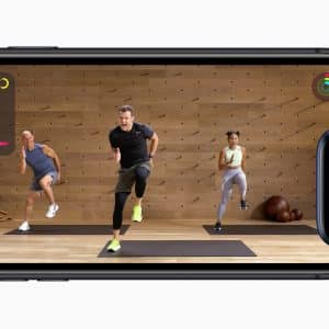 An iPhone displays a workout video with an instructor and two participants performing exercises in a wooden studio. Next to the iPhone, an Apple Watch shows matching workout metrics, including time, active calories burned, total calories, and heart rate.