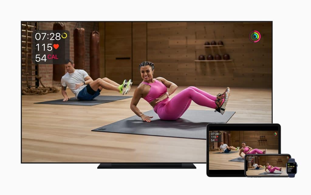 A TV screen, tablet, and smartwatch show a workout session featuring an instructor and a participant performing exercises on mats in a fitness studio. Utilizing Apple Fitness+, the screens display workout metrics like time, heart rate, and calories burned, seamlessly integrating with Apple services.