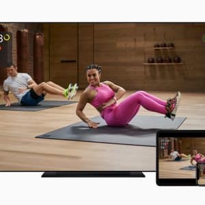 A TV screen, tablet, and smartwatch show a workout session featuring an instructor and a participant performing exercises on mats in a fitness studio. Utilizing Apple Fitness+, the screens display workout metrics like time, heart rate, and calories burned, seamlessly integrating with Apple services.