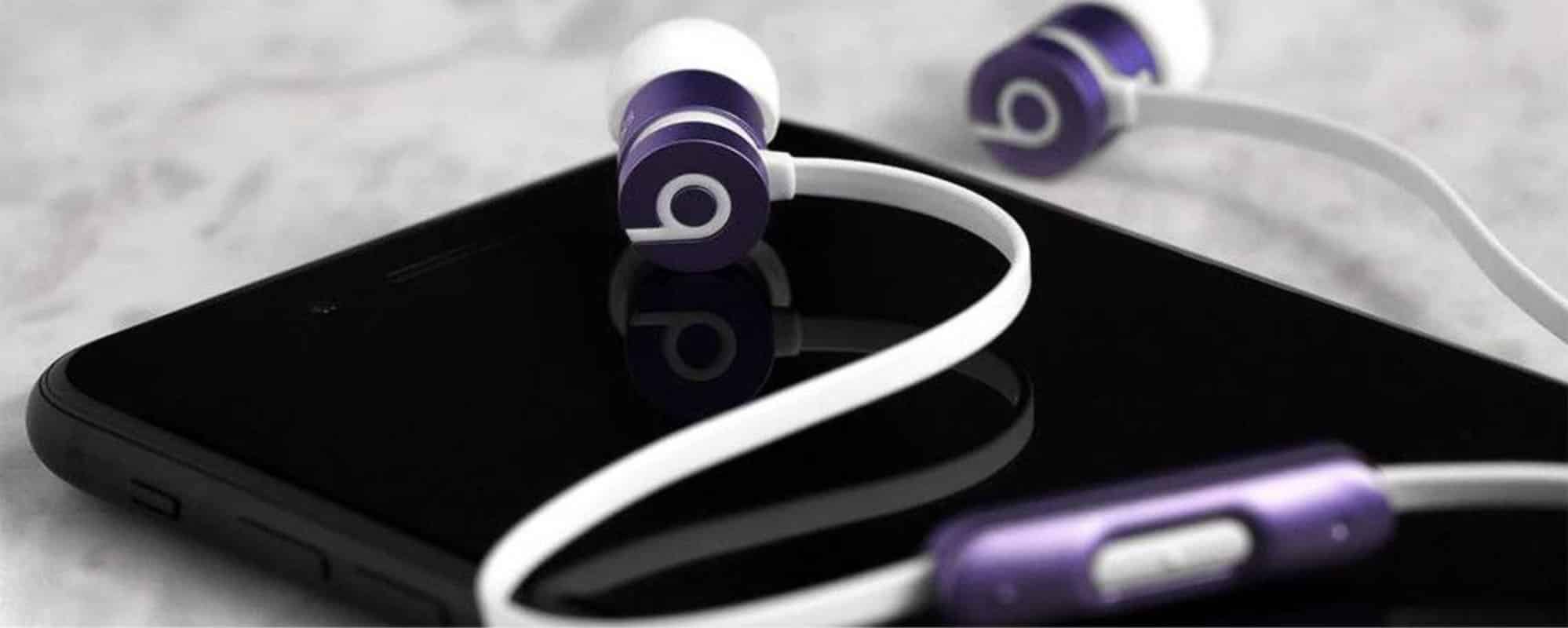 Close-up of a pair of purple and white in-ear Beats earphones—the best wired headphones— with their wire laying over a black smartphone. The earphones and phone sit on a marbled surface. The Beats logo is visible on the earphone buds.