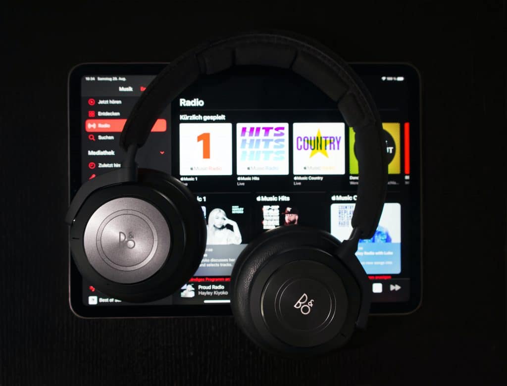 A pair of Beoplay headphones rests on an Apple iPad displaying the Apple Music streaming app. The app interface shows various radio stations and playlists with colorful covers against a dark background. The headphones are black with the Beoplay logo visible on the ear cups, adding a touch of elegance.