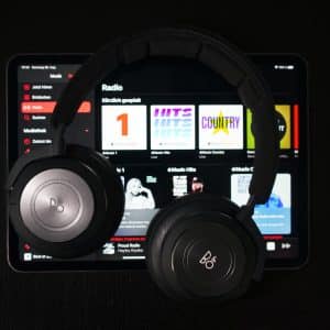 A pair of Beoplay headphones rests on an Apple iPad displaying the Apple Music streaming app. The app interface shows various radio stations and playlists with colorful covers against a dark background. The headphones are black with the Beoplay logo visible on the ear cups, adding a touch of elegance.