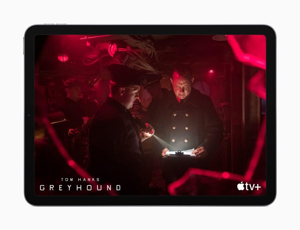 A scene from the movie "Greyhound" shown on a tablet screen. Two men in naval uniforms are in a dimly lit ship's interior; one holds a flashlight, while the other reads a document illuminated by the light. The background is illuminated by red lights, creating a tense atmosphere. "Tom Hanks Greyhound" and the Apple TV+ logo are displayed in the bottom corners.