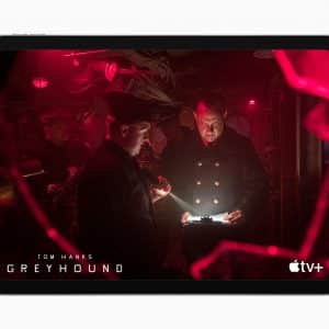 A scene from the movie "Greyhound" shown on a tablet screen. Two men in naval uniforms are in a dimly lit ship's interior; one holds a flashlight, while the other reads a document illuminated by the light. The background is illuminated by red lights, creating a tense atmosphere. "Tom Hanks Greyhound" and the Apple TV+ logo are displayed in the bottom corners.