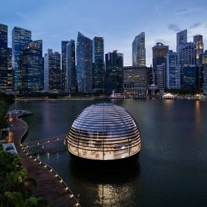 A futuristic spherical building with horizontal light strips floats in a waterfront area, reminiscent of Apple's innovative style. Surrounded by a boardwalk, the scene evokes the grandeur of Marina Bay Sands. The backdrop features a skyline of modern skyscrapers under a twilight sky with a few clouds.