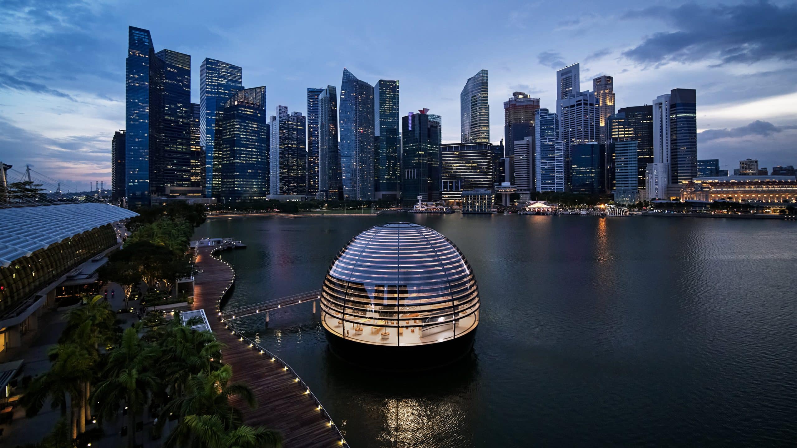 A futuristic spherical building with horizontal light strips floats in a waterfront area, reminiscent of Apple's innovative style. Surrounded by a boardwalk, the scene evokes the grandeur of Marina Bay Sands. The backdrop features a skyline of modern skyscrapers under a twilight sky with a few clouds.