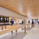 Spacious, modern Apple retail store with a wooden ceiling and long wooden tables displaying assorted electronic devices. Employees in blue shirts assist customers, and shelves with products line the walls. Located in Marina Bay Sands, the back of the store includes a staircase.