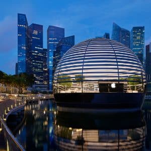 A floating, spherical Apple retail store with a lattice-like design is illuminated at dusk, sitting on the waters of Marina Bay Sands with numerous modern skyscrapers in the backdrop. People are visible on the boardwalk, taking photos and enjoying the view. Palms and greenery adorn the area.