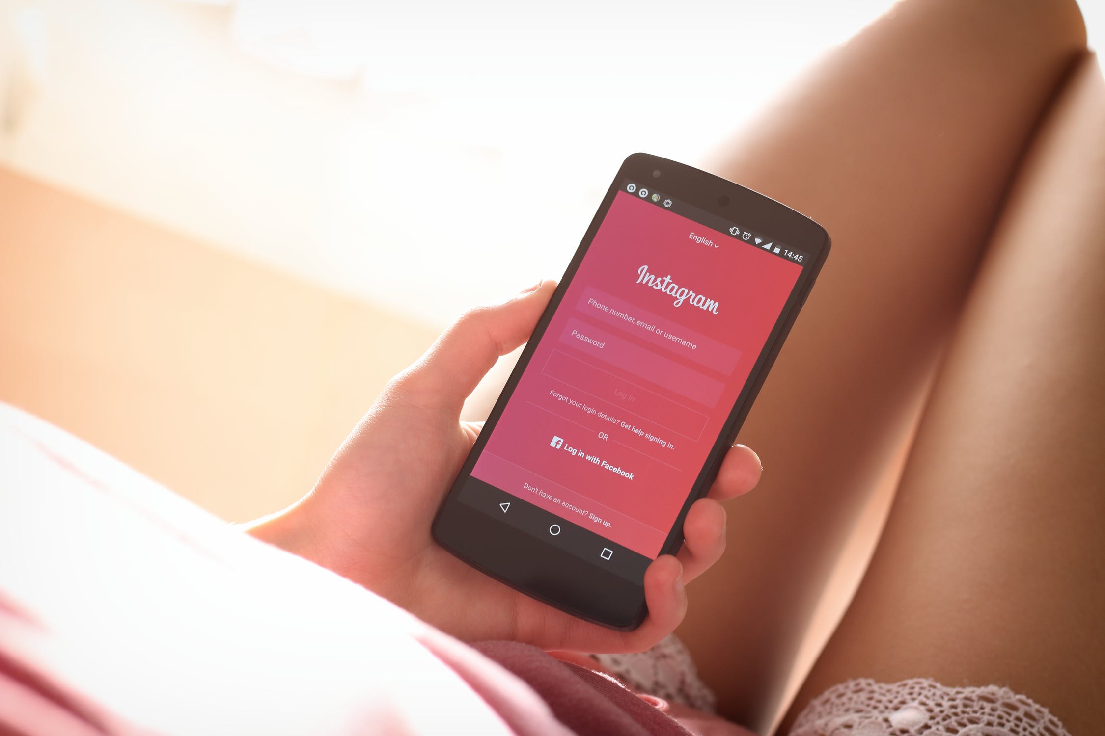 A person holding a smartphone displaying the Instagram login screen, which is glowing with a bright pink background. They are sitting with their legs comfortably bent, wearing lace-edged shorts, perhaps ready to dive into an Instagram marketing guide for successful marketing.