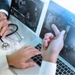 Two medical professionals discuss something displayed on a laptop. One person points at the screen, while the other uses a computer mouse. A stethoscope and another screen displaying medical charts are visible in the background, highlighting the evolving technology in the healthcare industry.