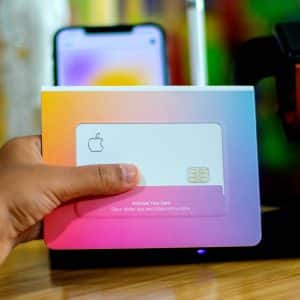 A hand holding an Apple Card in its packaging with an iPhone displaying the card activation setup screen in the background. A black Apple Watch with a brown band is on a charging stand next to the iPhone on a wooden surface, marking the Apple Card launch in other countries.