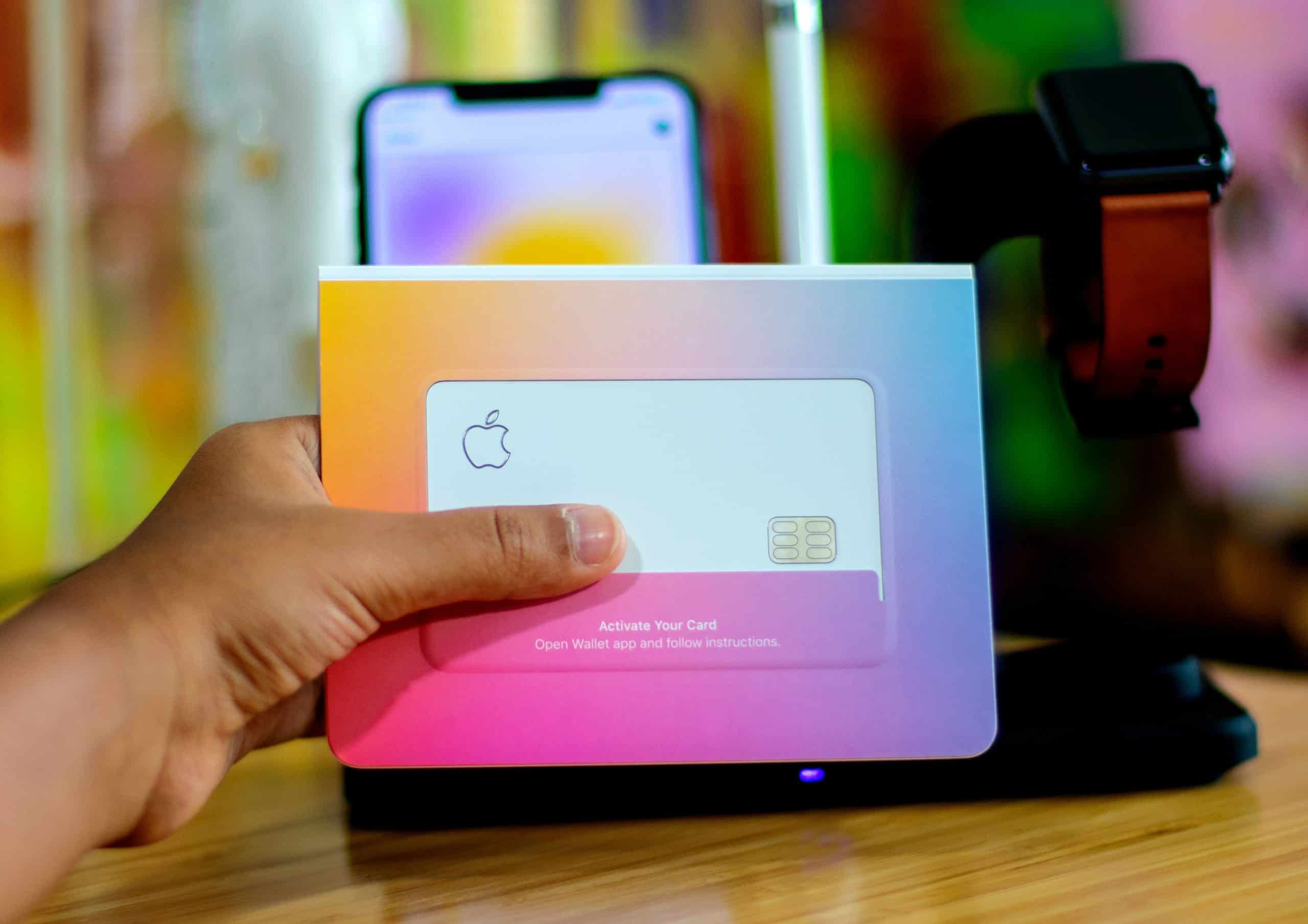 A hand holding an Apple Card in its packaging with an iPhone displaying the card activation setup screen in the background. A black Apple Watch with a brown band is on a charging stand next to the iPhone on a wooden surface, marking the Apple Card launch in other countries.