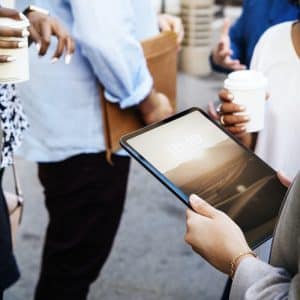 A group of people are standing and conversing outdoors. One person is holding an Apple iPad displaying the time "10:10," while others hold coffee cups. They are dressed in business casual attire. The background is slightly blurred, focusing on the interaction and their environmentally-friendly surroundings.