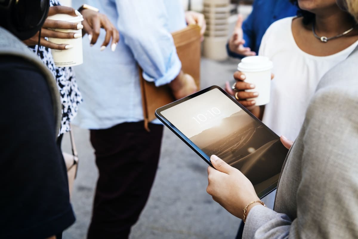 A group of people are standing and conversing outdoors. One person is holding an Apple iPad displaying the time "10:10," while others hold coffee cups. They are dressed in business casual attire. The background is slightly blurred, focusing on the interaction and their environmentally-friendly surroundings.