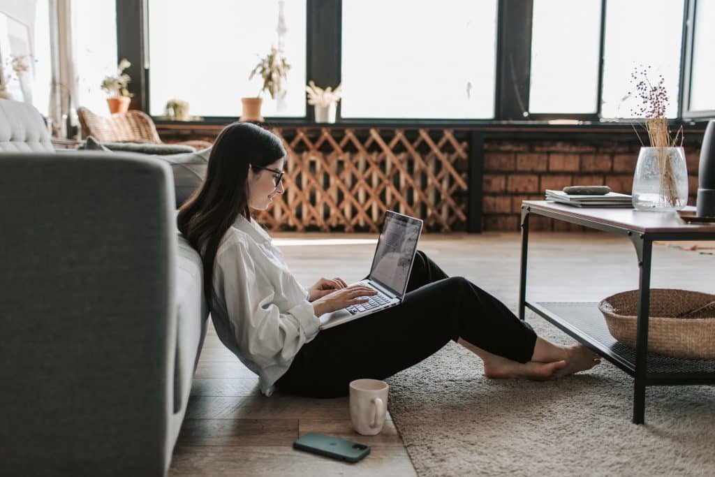 A woman with long hair and glasses is sitting on the floor, leaning against a couch, and typing on a laptop. She is in a well-lit living room with large windows, plants, a wooden table, and a mug and smartphone placed on the floor next to her. Her Mac VPN software from 2020 ensures her privacy as she works.