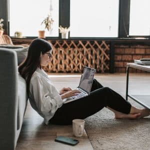 A woman with long hair and glasses is sitting on the floor, leaning against a couch, and typing on a laptop. She is in a well-lit living room with large windows, plants, a wooden table, and a mug and smartphone placed on the floor next to her. Her Mac VPN software from 2020 ensures her privacy as she works.