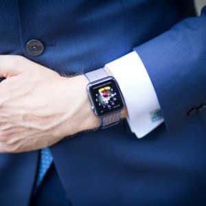 A person wearing a formal blue suit with a light blue tie has their left hand resting on their stomach. They are wearing an Apple Watch on their left wrist, displaying a colorful screen with the ECG feature active. The suit sleeve is rolled up slightly, exposing the watch.