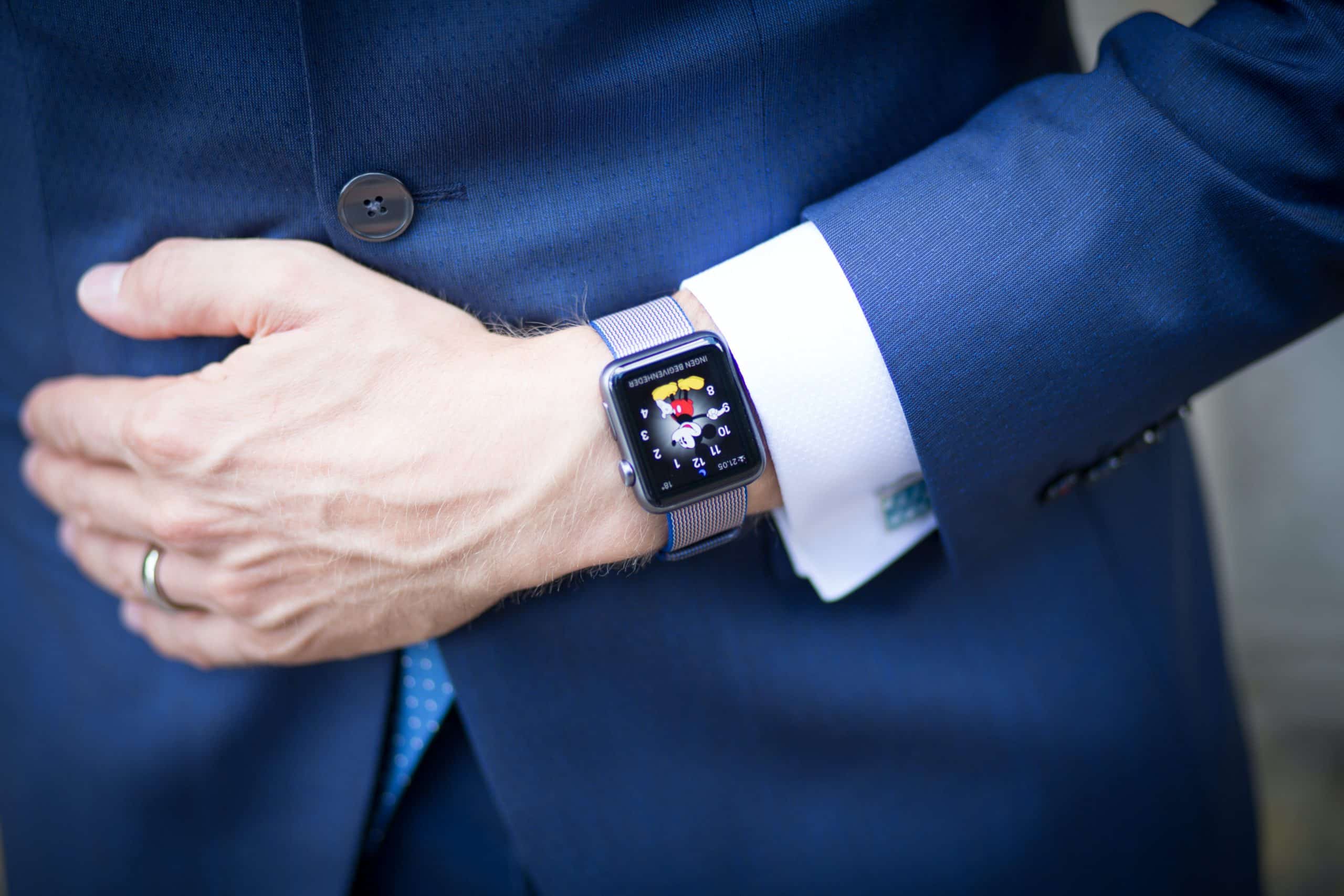 A person wearing a formal blue suit with a light blue tie has their left hand resting on their stomach. They are wearing an Apple Watch on their left wrist, displaying a colorful screen with the ECG feature active. The suit sleeve is rolled up slightly, exposing the watch.