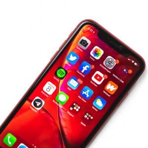 A red Apple smartphone with a vibrant screen displaying various app icons on a red-themed background. Apps include Photos, Calendar, Facebook, Twitter, Spotify, Maps, Settings, and others. The device is angled slightly to the right on a white surface.