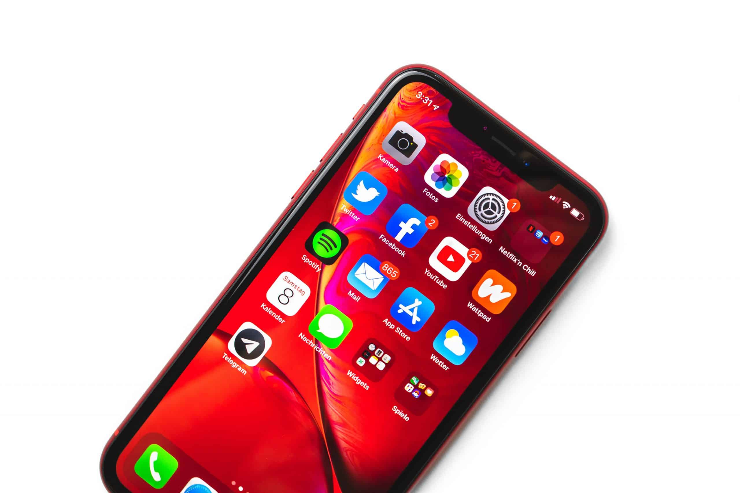 A red Apple smartphone with a vibrant screen displaying various app icons on a red-themed background. Apps include Photos, Calendar, Facebook, Twitter, Spotify, Maps, Settings, and others. The device is angled slightly to the right on a white surface.