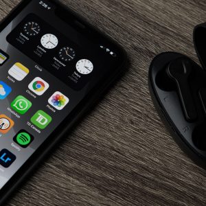 A sleek Apple smartphone displaying various app icons, including a 2021 ad privacy feature, rests on a wooden surface next to an open black case containing wireless earbuds. The phone screen shows clocks, weather, and other app icons.