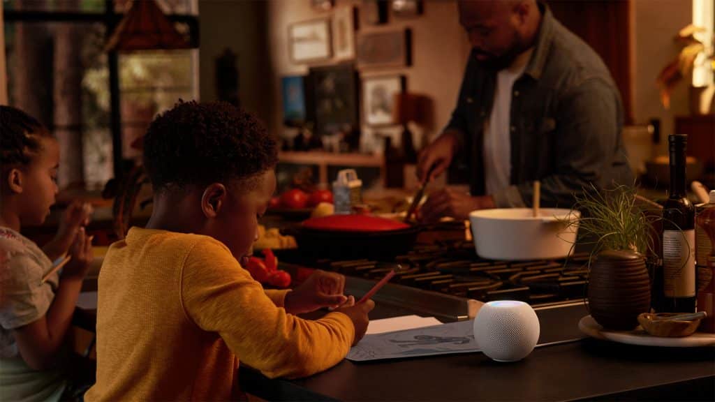 A child is drawing at a kitchen counter while another child sits beside them. In the background, an adult is chopping vegetables. On the counter near the children, there is a low-cost smart speaker, a small potted plant, and various kitchen items.