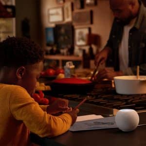 A child is drawing at a kitchen counter while another child sits beside them. In the background, an adult is chopping vegetables. On the counter near the children, there is a low-cost smart speaker, a small potted plant, and various kitchen items.