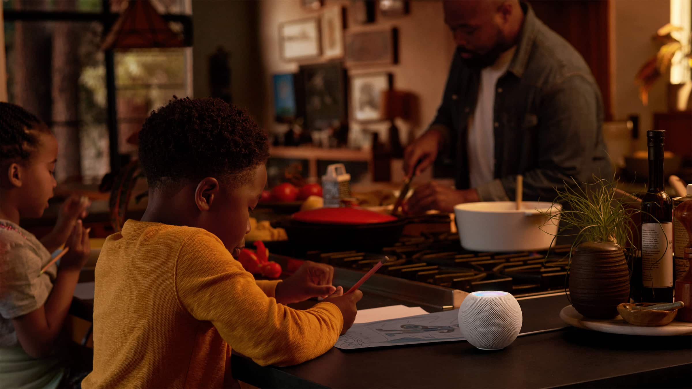 A child is drawing at a kitchen counter while another child sits beside them. In the background, an adult is chopping vegetables. On the counter near the children, there is a low-cost smart speaker, a small potted plant, and various kitchen items.