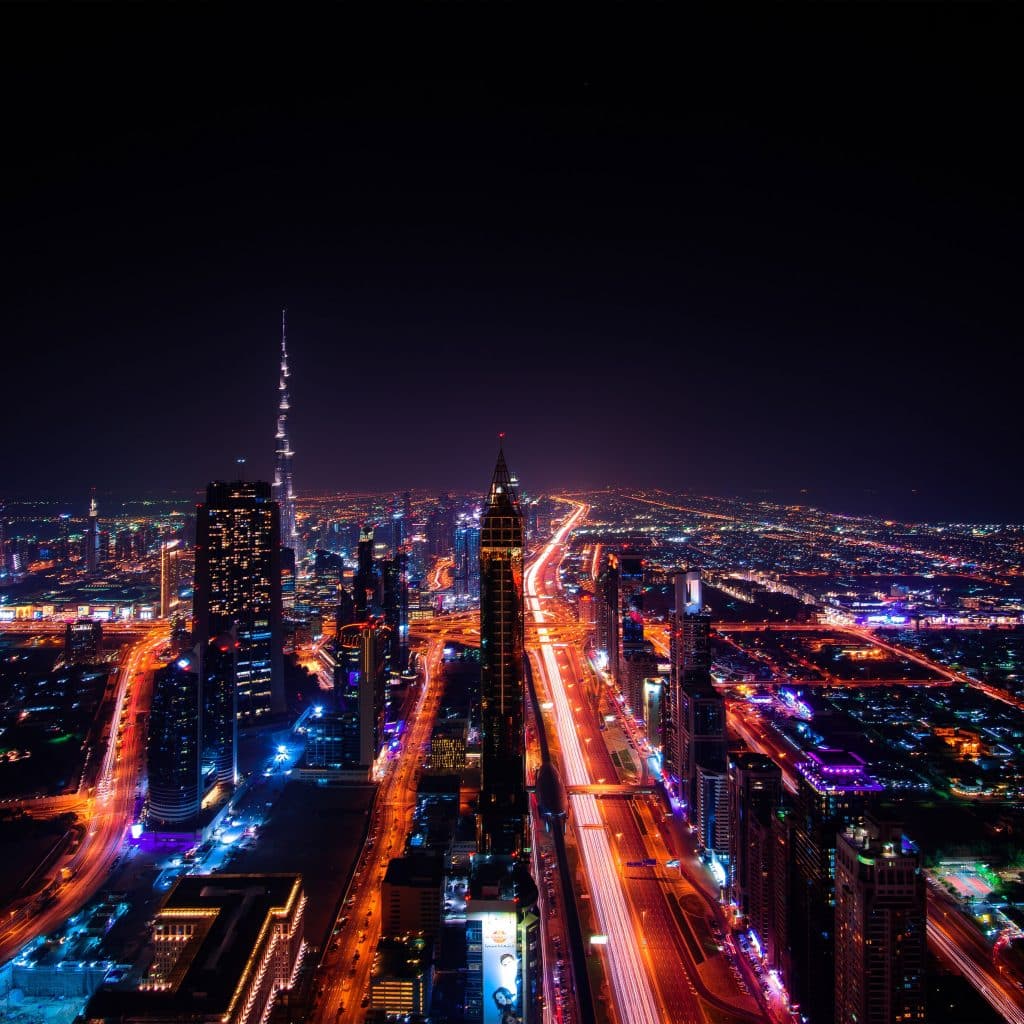 A nighttime aerial view of a vibrant cityscape illuminated with colorful lights. Skyscrapers, housing numerous companies, line the roads, and a prominent tower stands illuminated in the distance. Streets are aglow with traffic and streetlights, creating a dynamic and lively urban scene reflecting up-to-date solutions.