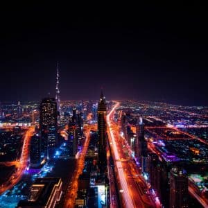 A nighttime aerial view of a vibrant cityscape illuminated with colorful lights. Skyscrapers, housing numerous companies, line the roads, and a prominent tower stands illuminated in the distance. Streets are aglow with traffic and streetlights, creating a dynamic and lively urban scene reflecting up-to-date solutions.