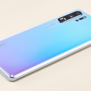 A sleek, silver Huawei smartphone with a gradient of blue and purple on its back featuring a vertical triple-camera setup and a flash. As one of the biggest smartphone players, Huawei's device is placed on a light beige surface, showcasing its rear design and reflective metallic edges.