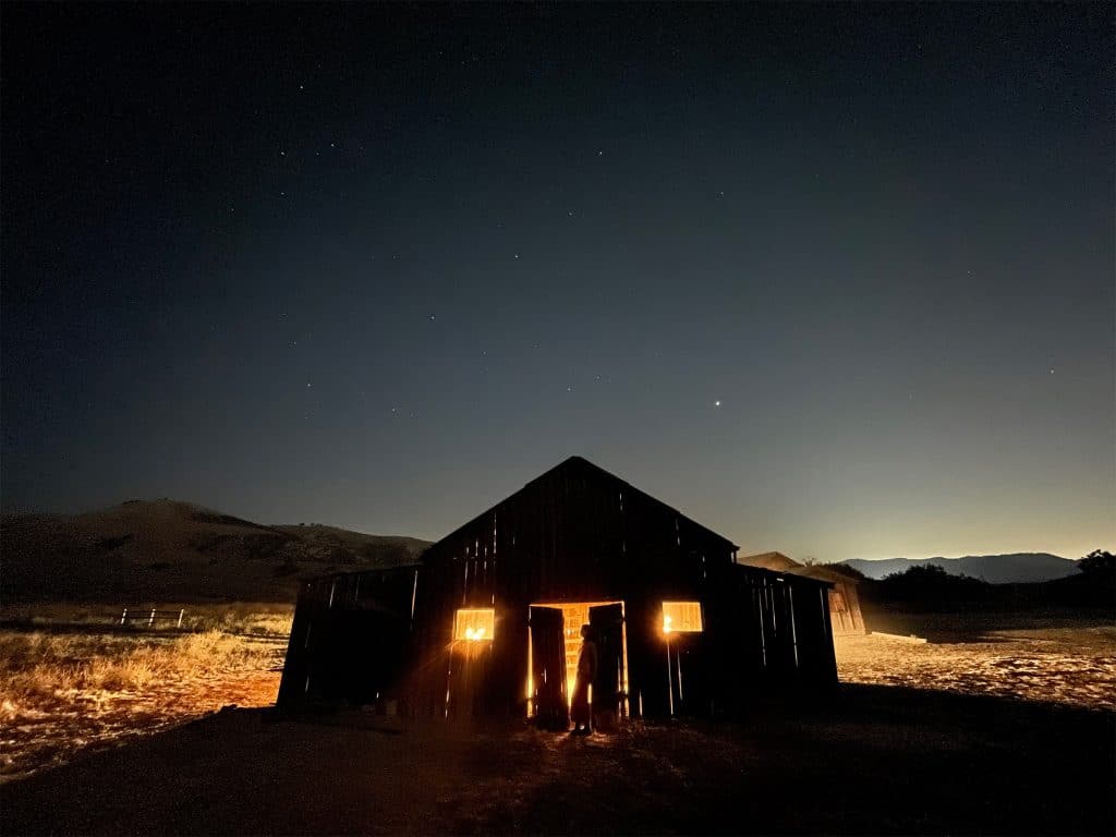 A dimly illuminated barn stands in a dark, open area under a clear night sky dotted with stars. Soft, warm light seeps through the open doors and windows, casting a faint glow on the surrounding ground. A person is silhouetted in the doorway.