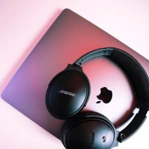 Black Bose over-ear headphones resting on a closed silver Apple MacBook. The softly lit background features shades of pink and purple, accentuating the glowing Apple logo in the middle of the MacBook.