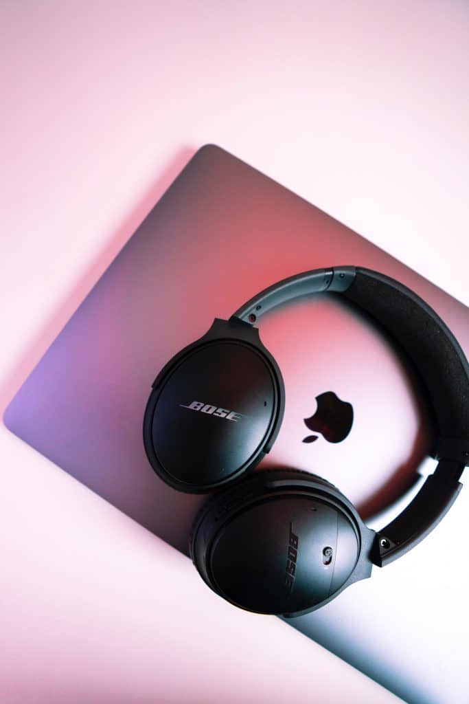 Black Bose over-ear headphones resting on a closed silver Apple MacBook. The softly lit background features shades of pink and purple, accentuating the glowing Apple logo in the middle of the MacBook.