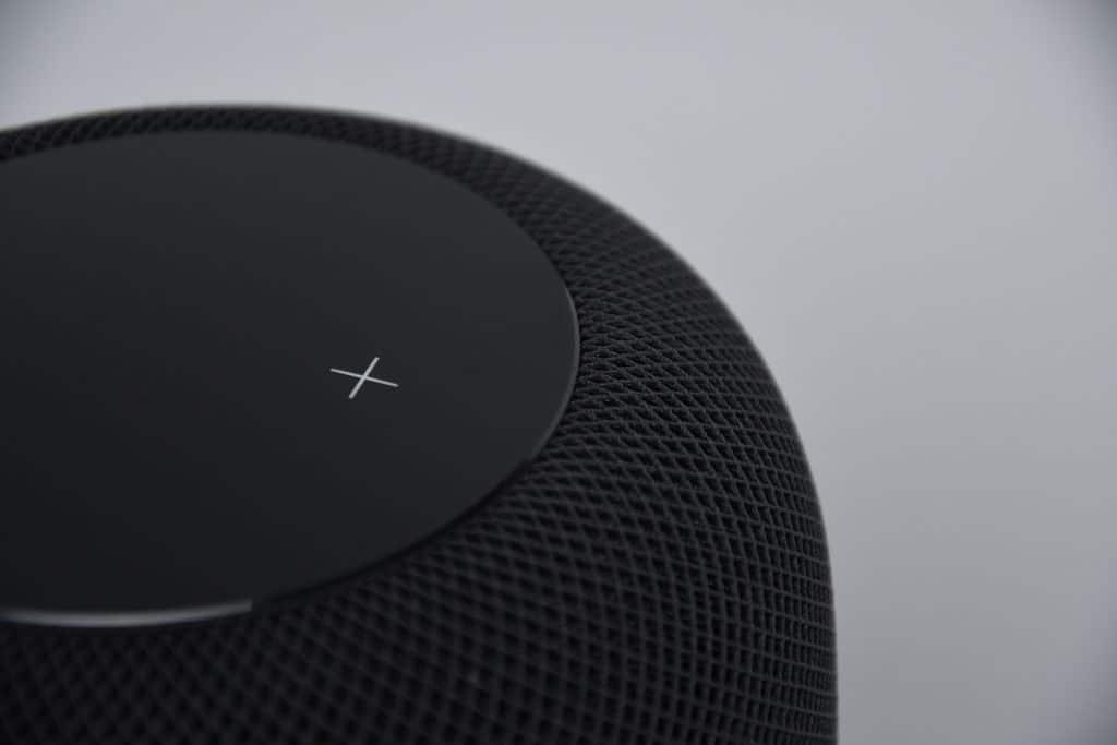 A close-up of an Apple HomePod Mini with a rounded, mesh-like exterior and a smooth top surface that displays a white "X" symbol. The background is plain and gray.