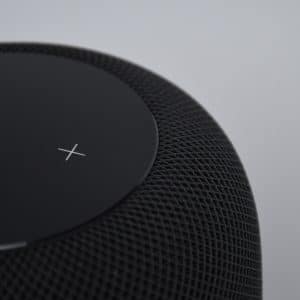 A close-up of an Apple HomePod Mini with a rounded, mesh-like exterior and a smooth top surface that displays a white "X" symbol. The background is plain and gray.