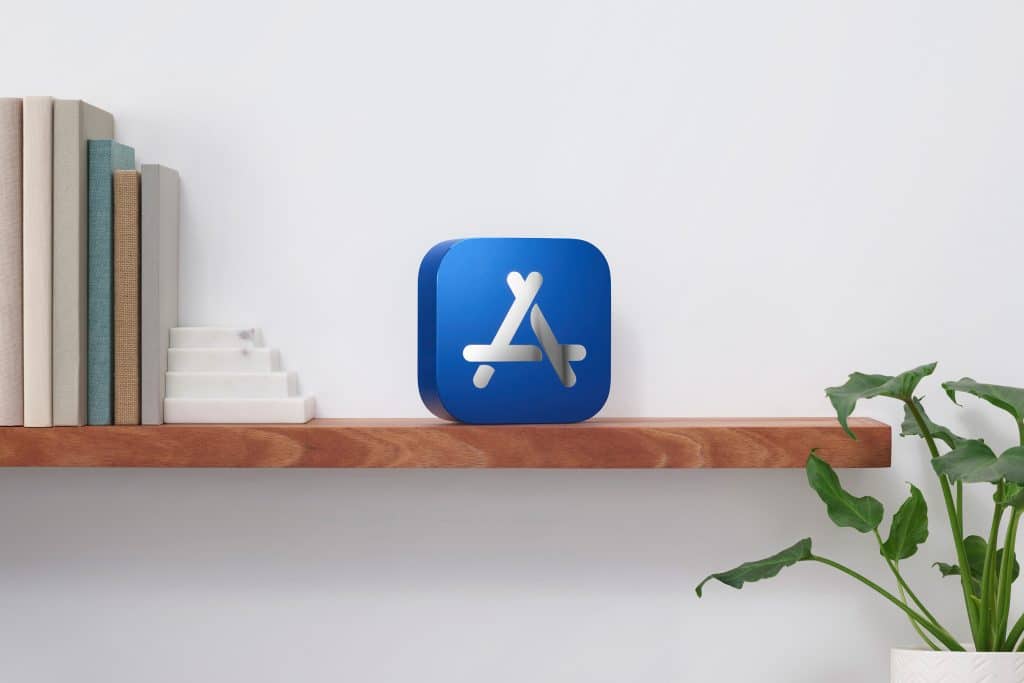 A wooden shelf against a white wall holds several books, a small staircase-like marble ornament, and a decorative object in the shape of the App Store logo, celebrating its Best of 2020 iPhone apps. A leafy green plant is situated on a table to the right of the shelf.