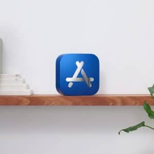 A wooden shelf against a white wall holds several books, a small staircase-like marble ornament, and a decorative object in the shape of the App Store logo, celebrating its Best of 2020 iPhone apps. A leafy green plant is situated on a table to the right of the shelf.