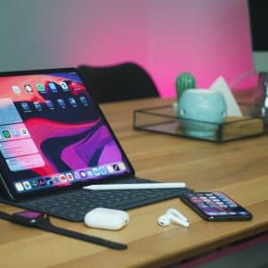 A tablet with a keyboard and stylus is placed on a wooden desk. Nearby, there are earbuds, a smartwatch, a smartphone, and a charging case. In the background, there is a shelf with decorative items and an iMac running macOS. The scene is illuminated with a pink light.