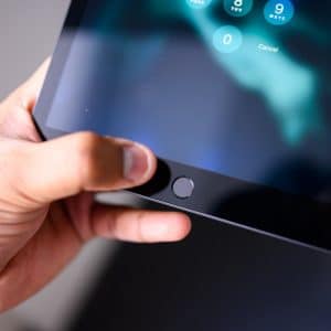 A hand holds an iPad, with the thumb pressing the home button on the front. The tablet screen displays a numeric keypad for entering a password, guiding the user through secure access.