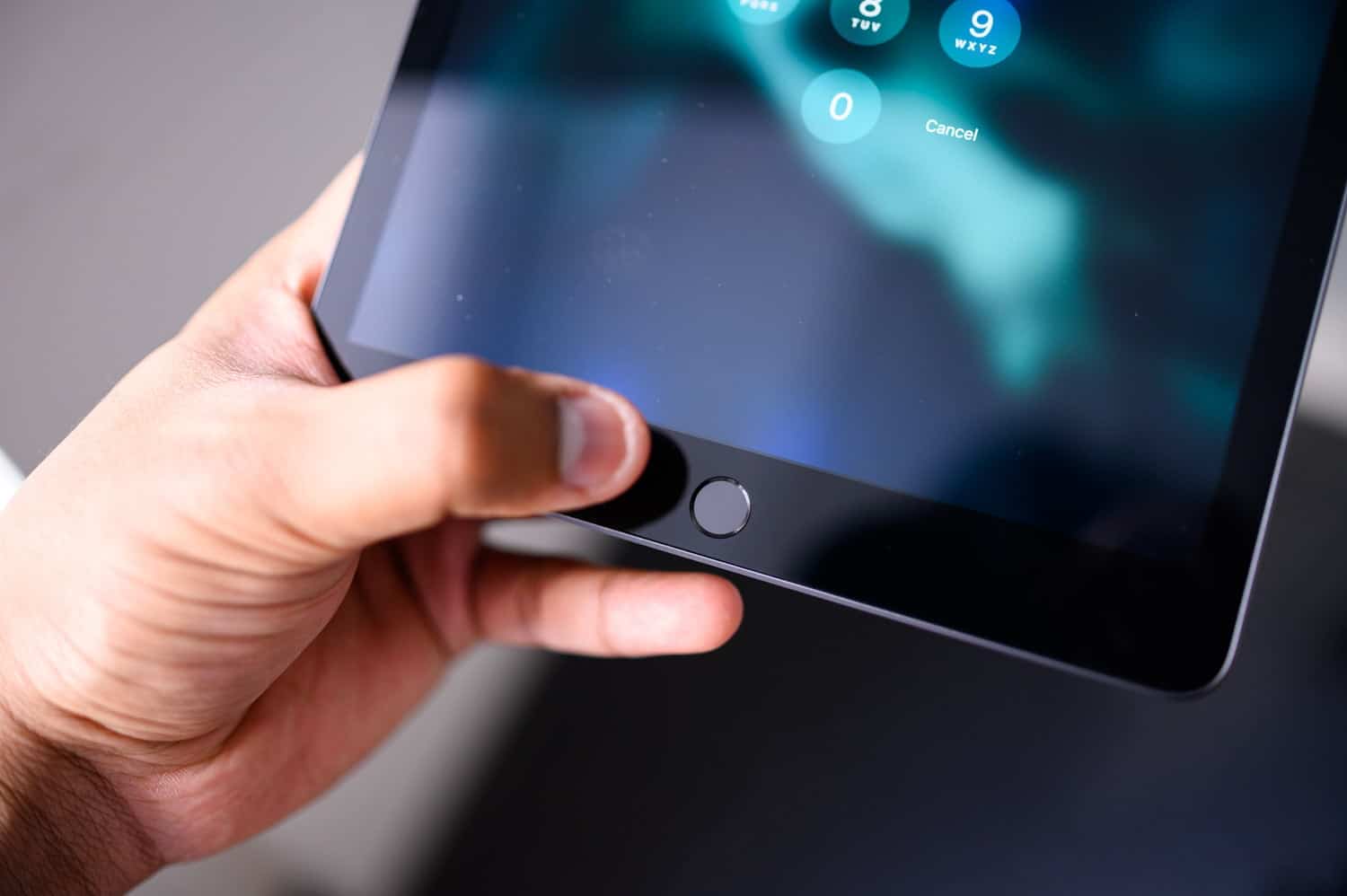 A hand holds an iPad, with the thumb pressing the home button on the front. The tablet screen displays a numeric keypad for entering a password, guiding the user through secure access.