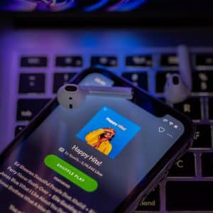 A smartphone displaying a music streaming app with the playlist "Happy Hits!" is placed on a laptop keyboard. White wireless earbuds lie on the phone. The background glows with purple and blue lights, showcasing Spotify's new ad-supported streaming tier.