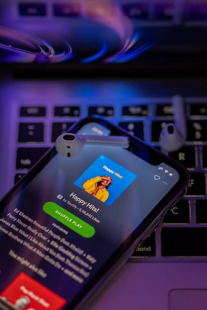 A smartphone displaying a music streaming app with the playlist "Happy Hits!" is placed on a laptop keyboard. White wireless earbuds lie on the phone. The background glows with purple and blue lights, showcasing Spotify's new ad-supported streaming tier.