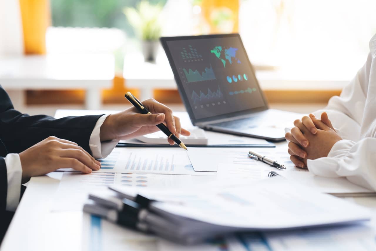 Two individuals are engaged in a business discussion at a desk filled with documents and charts. One person, aiming to explain the purpose of their strategy, is pointing at a chart with a pen, while a laptop displaying financial graphs and world maps is open in the background.