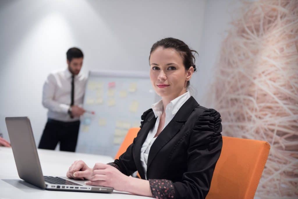 A woman in a black blazer is seated at a table, working on her laptop with a smile of understanding. In the background, a man in a white shirt and tie stands purposefully in front of a whiteboard with sticky notes. The setting appears to be an office or meeting room.