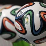 Close-up image of Adidas Brazuca soccer balls, the official match balls of the 2014 FIFA World Cup. They feature a white base with colorful swirling patterns in blue, orange, and green, showcasing the tournament logo and Adidas branding—highlights seen on TV rights broadcasts of major events like the FIFA Club World Cup.
