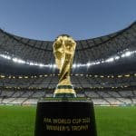 Image of the FIFA World Cup 2022 Winner's Trophy displayed in an empty stadium. The gold trophy stands prominently on a black pedestal against the backdrop of the stadium's seating and roof structure, showcasing its intricate design under stadium lights, akin to those seen during FIFA Club World Cup broadcasts.