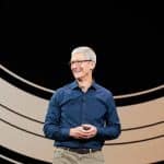 A person with short gray hair and glasses stands on a stage, smiling while wearing a dark blue shirt and khaki pants. The background features large, curved, concentric lines, setting the scene for the anticipated discussion on Q2 2024 analyst expectations for Apple earnings.