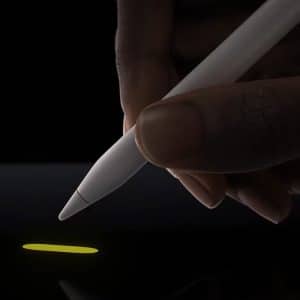 A hand holding a white Apple Pencil Pro is drawing a small yellow line on the latest iPads' touchscreen. The background is black, highlighting the illuminated line and stylus tip, showcasing the advanced features of this cutting-edge device.