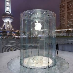 A cylindrical glass entrance featuring the Apple logo leads to an underground Apple Store in a cityscape with tall, illuminated buildings and the iconic Oriental Pearl Tower in the background at night. Amid a sales surge due to iPhone discounts in China, a few people are walking near the entrance.